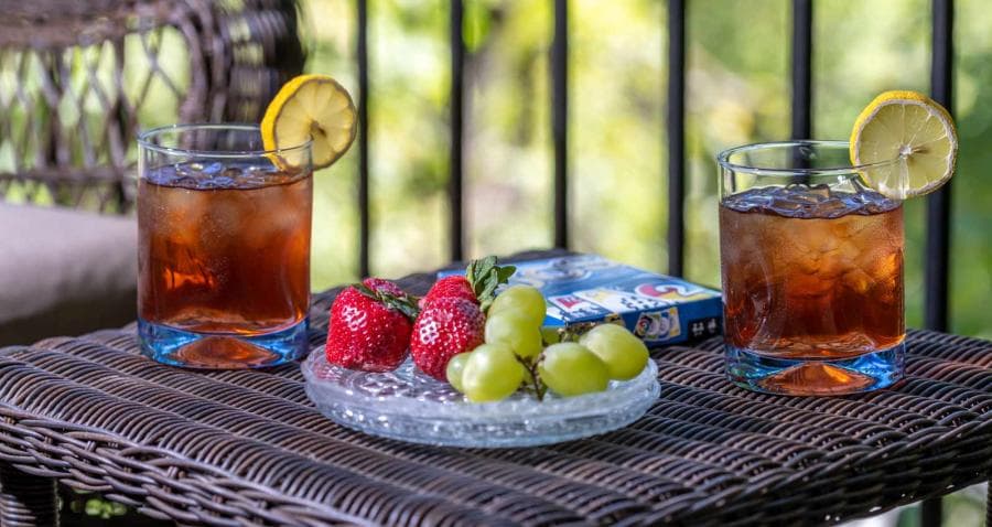 Two glasses of iced tea with lemon sit next to a plate of strawberries and green grapes on a wicker table.