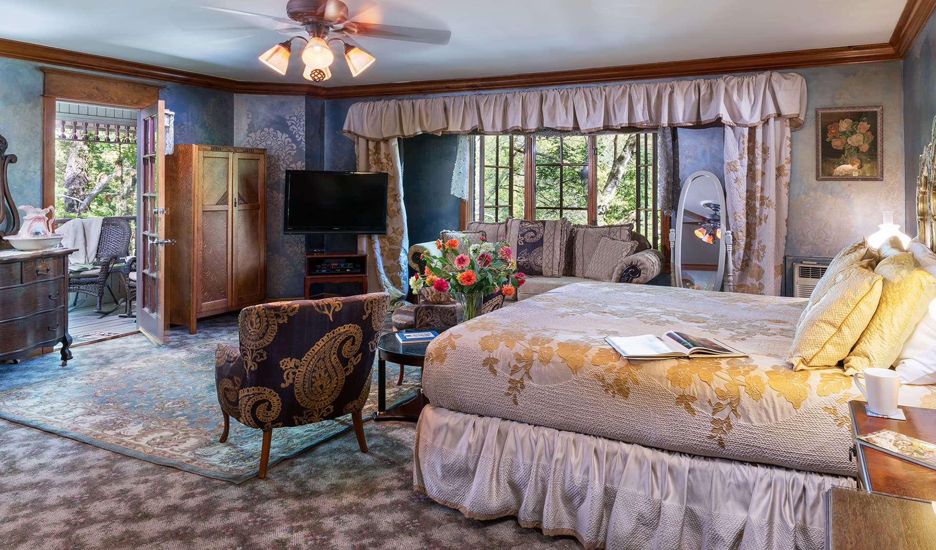A cozy, elegantly decorated bedroom with a television, comfortable seating, and a view of greenery through large windows.