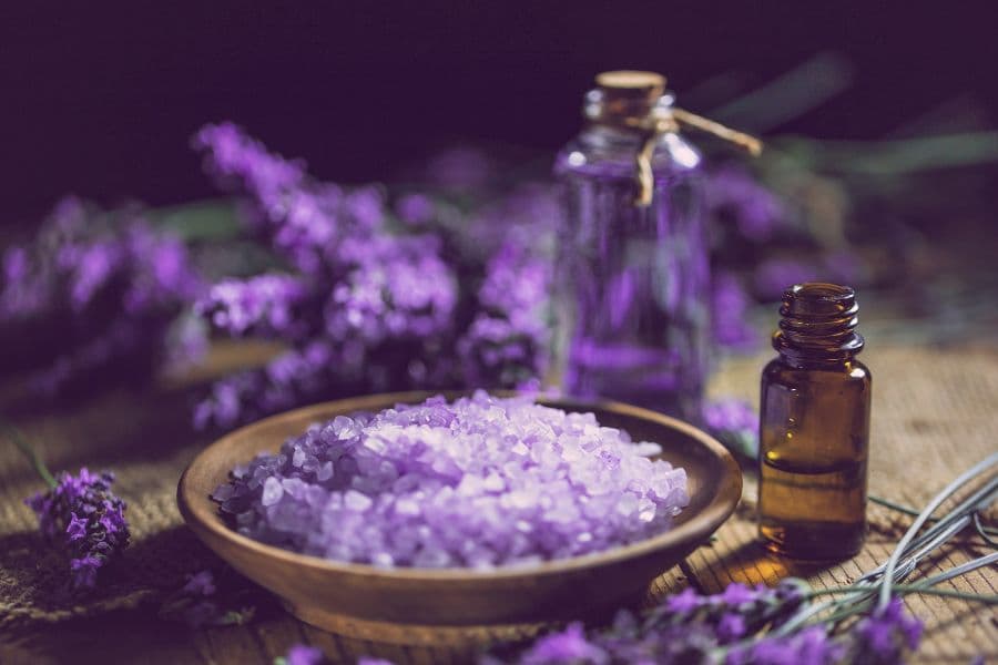 A wooden bowl filled with purple bath salts, accompanied by glass essential oil bottles and lavender sprigs.