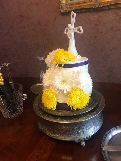 A two-tiered cake decorated with white and yellow flowers and topped with a cross, displayed on a silver platter.