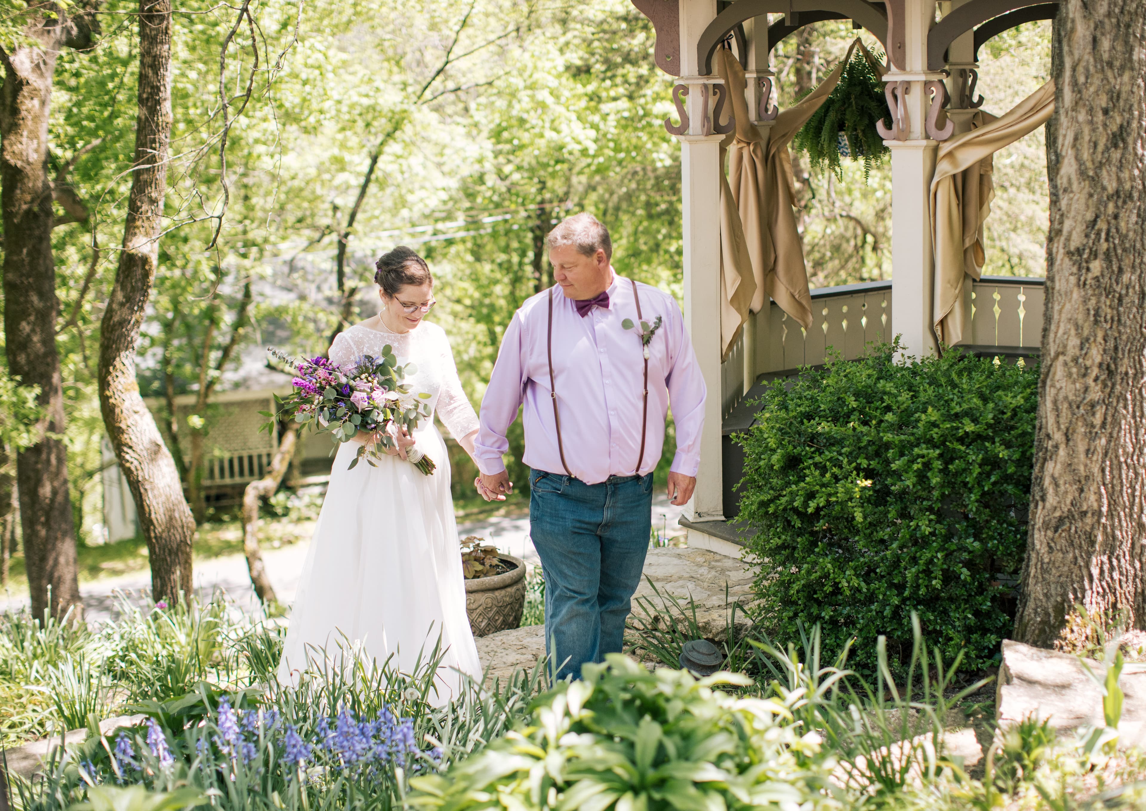 A bride and her father walk hand in hand through a lush garden toward a gazebo.