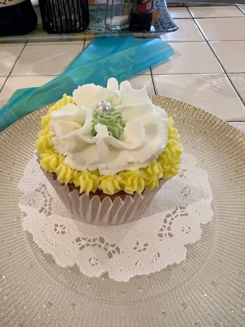 A decorated cupcake with yellow and white frosting topped with a green flower and a pearl, placed on a lace doily.