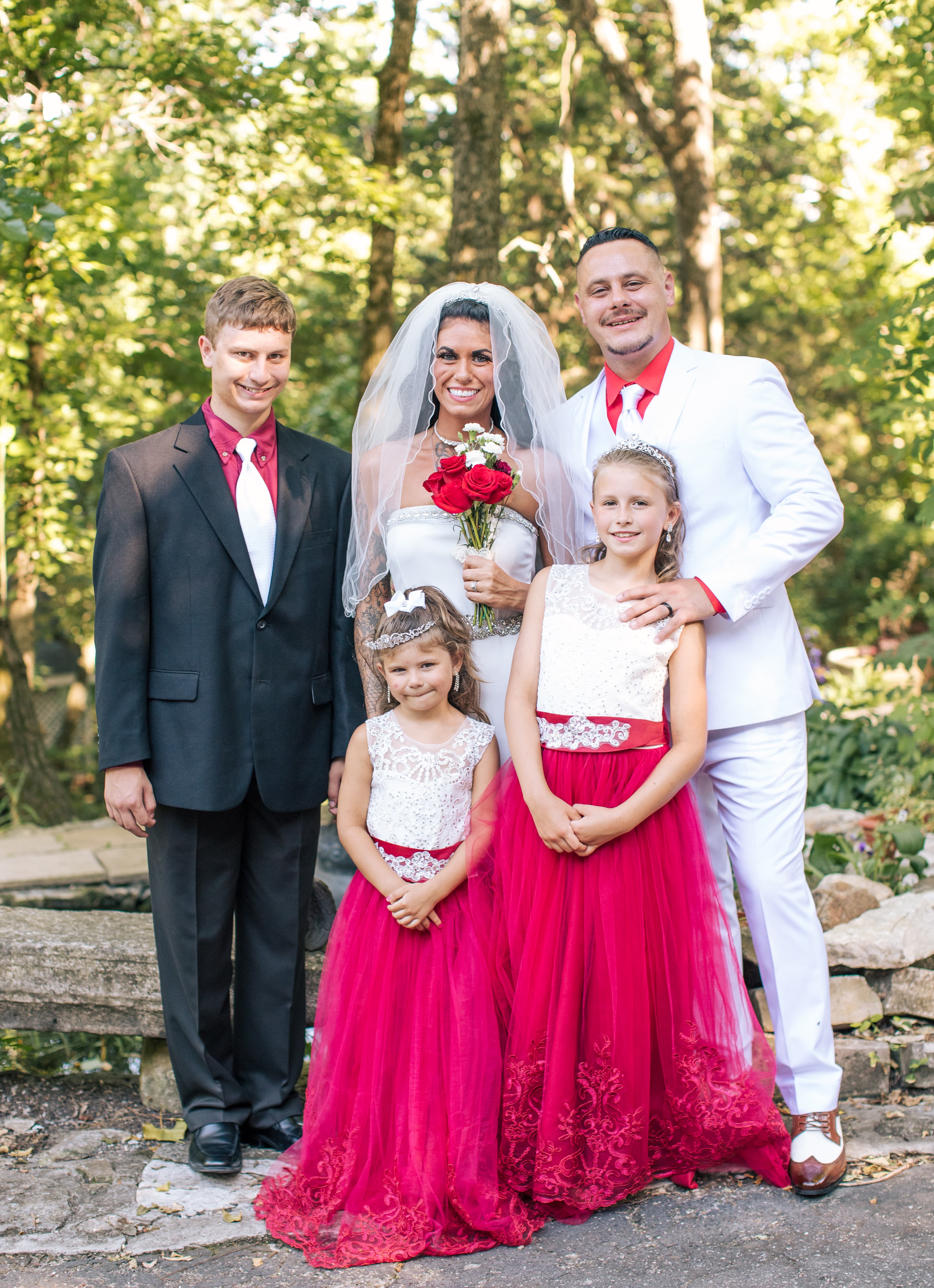 A bride and groom pose with three children in formal attire, surrounded by greenery.