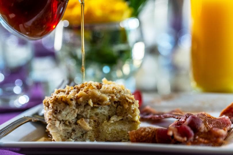 A close-up of a plate with a piece of baked dish being drizzled with syrup, alongside crispy bacon and a blurred background of drinks and flowers.