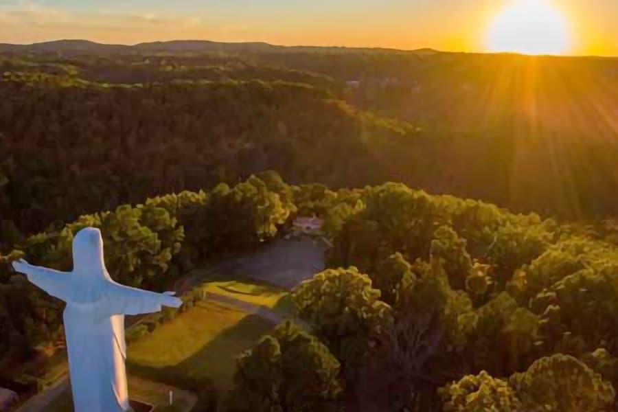 Aerial view of a large statue with arms outstretched against a sunset over a forested landscape.