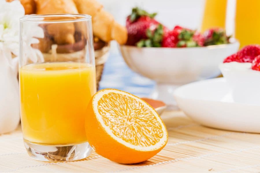 A glass of orange juice beside a halved orange, with a background of fruits and baked goods.