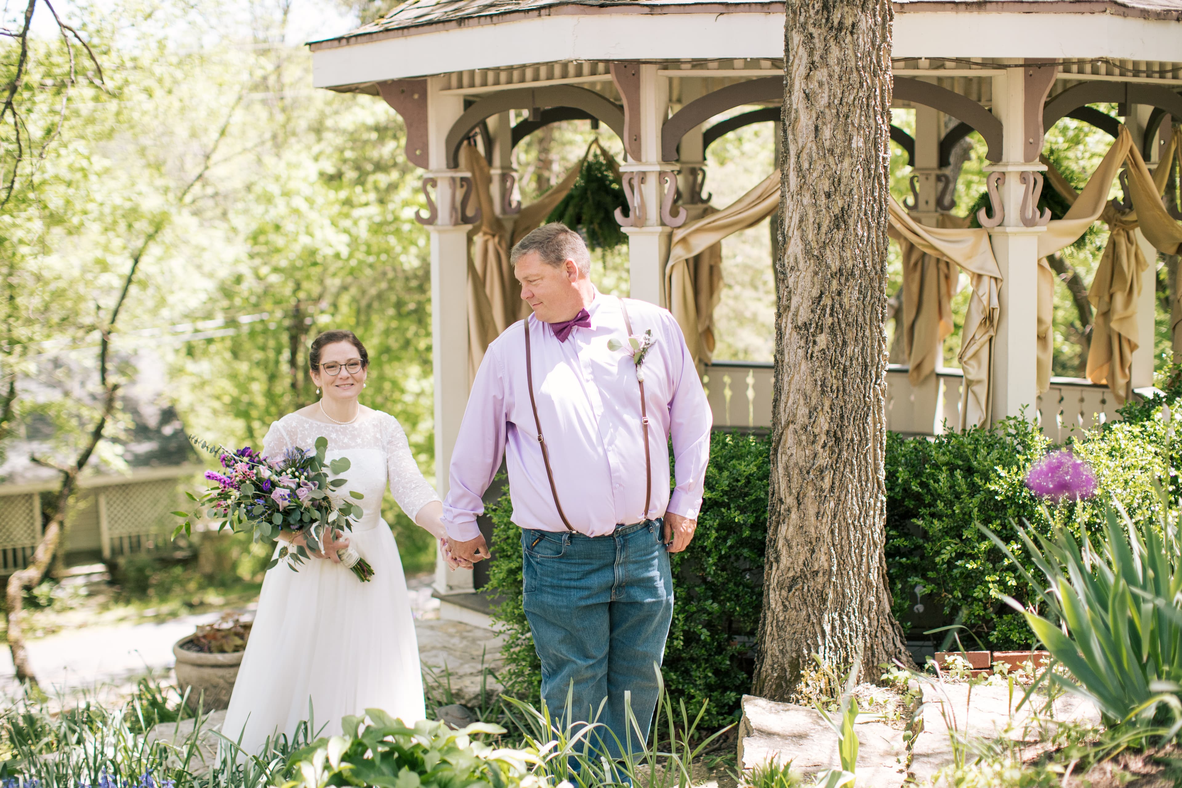 A bride and a man hold hands, walking together in a garden near a gazebo.