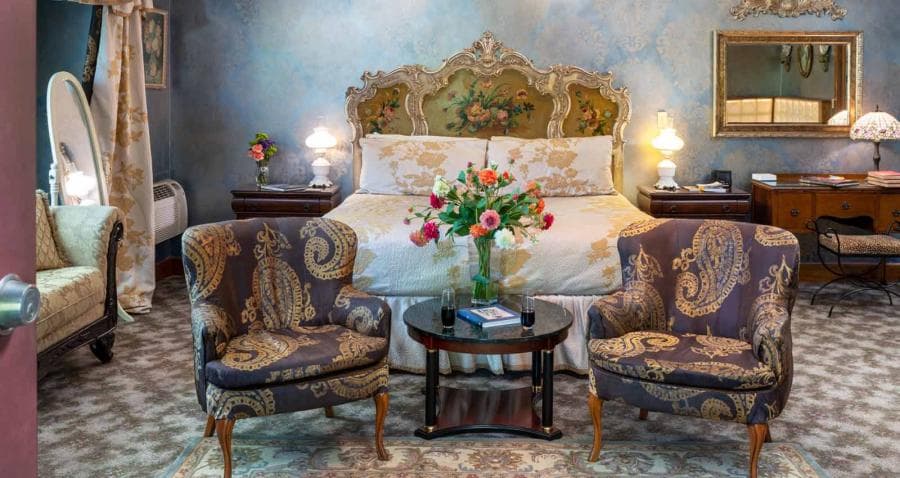 A cozy bedroom featuring a decorative bed, floral arrangement, two patterned armchairs, and elegant furnishings.