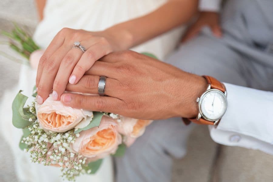 A close-up of two hands with wedding rings, resting on a bouquet of peach roses.