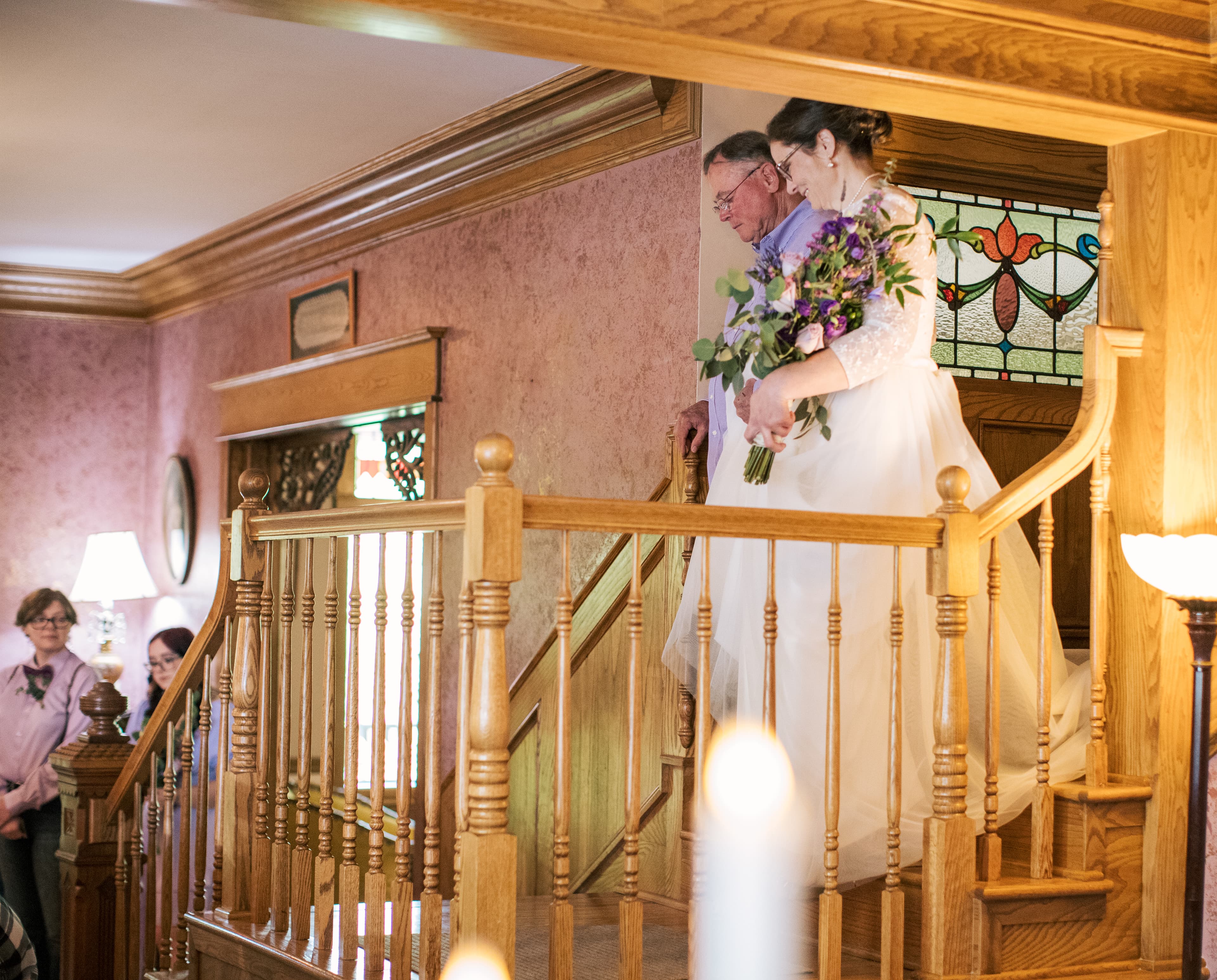 A bride descends a staircase with her father, surrounded by guests in a warmly lit interior.