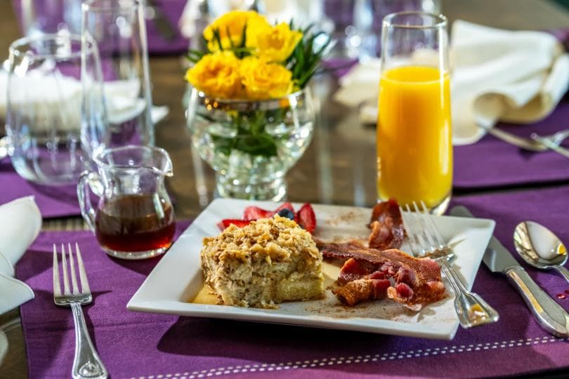 A plate with baked dish, bacon, strawberries, and syrup, accompanied by a glass of orange juice and a vase of yellow flowers on a purple tablecloth.
