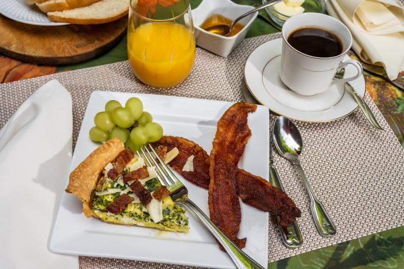 A breakfast plate featuring a slice of quiche, grapes, crispy bacon, a glass of orange juice, and a cup of black coffee.