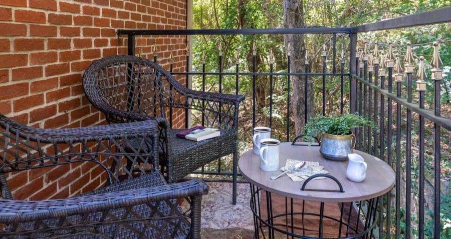 A cozy balcony with wicker chairs, a small table, coffee mugs, and greenery set against a backdrop of trees and brick walls.