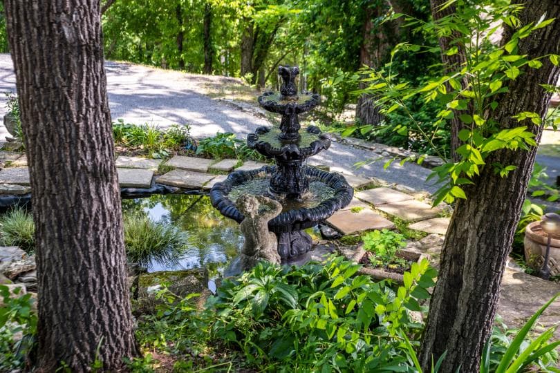 A black stone fountain surrounded by greenery and stepping stones in a serene garden setting.