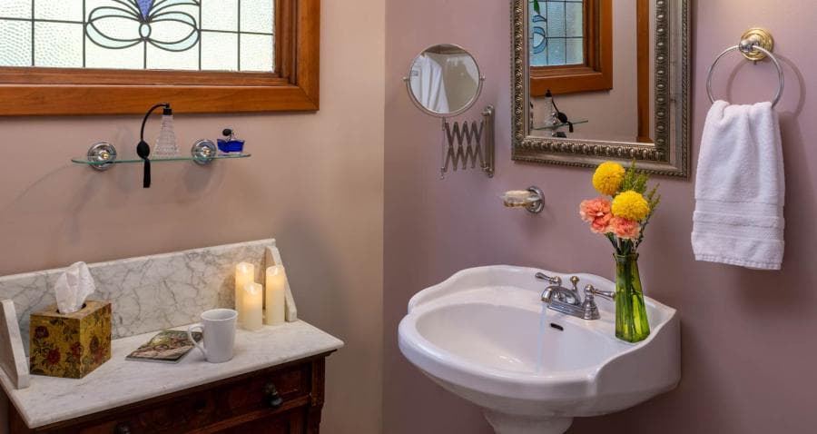 A cozy bathroom featuring a small sink, floral arrangement, candles, and a decorative window.