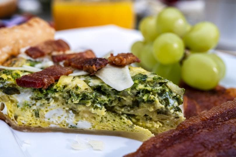 A plate featuring a sliced frittata topped with bacon, alongside green grapes and crispy bacon.