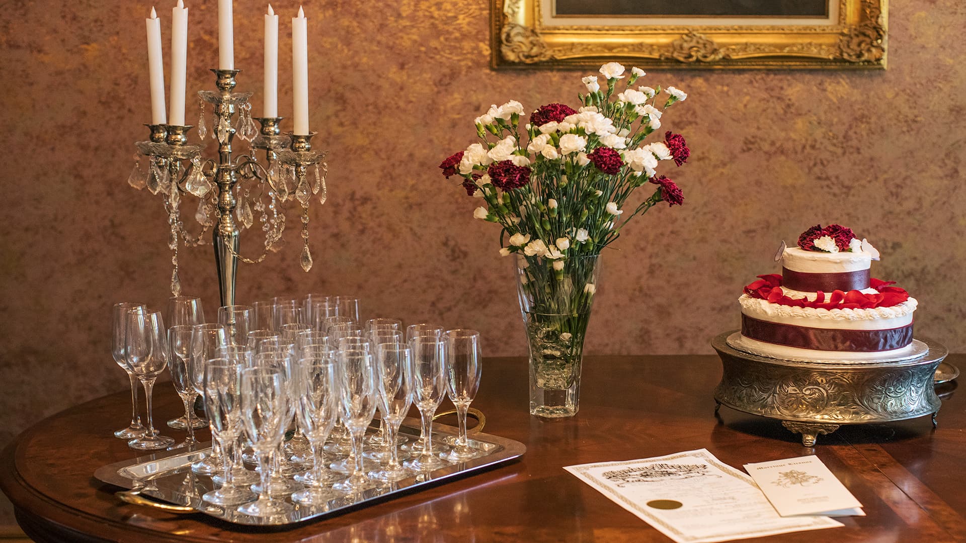 A decorative table setting featuring a silver candelabra, a bouquet of mixed flowers, champagne flutes, a wedding cake, and celebratory documents.