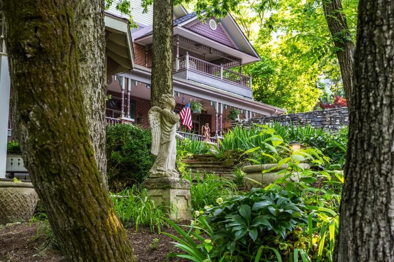 A lush garden with a decorative statue in the foreground and a house featuring a porch and an American flag in the background.