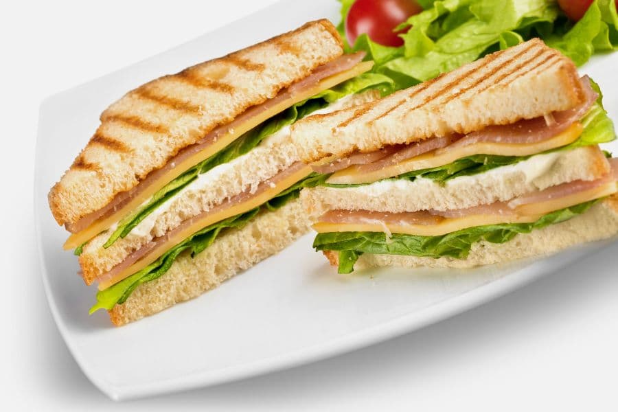 A grilled sandwich with layers of ham, cheese, lettuce, and cream cheese on toasted bread, served with a side of greens.