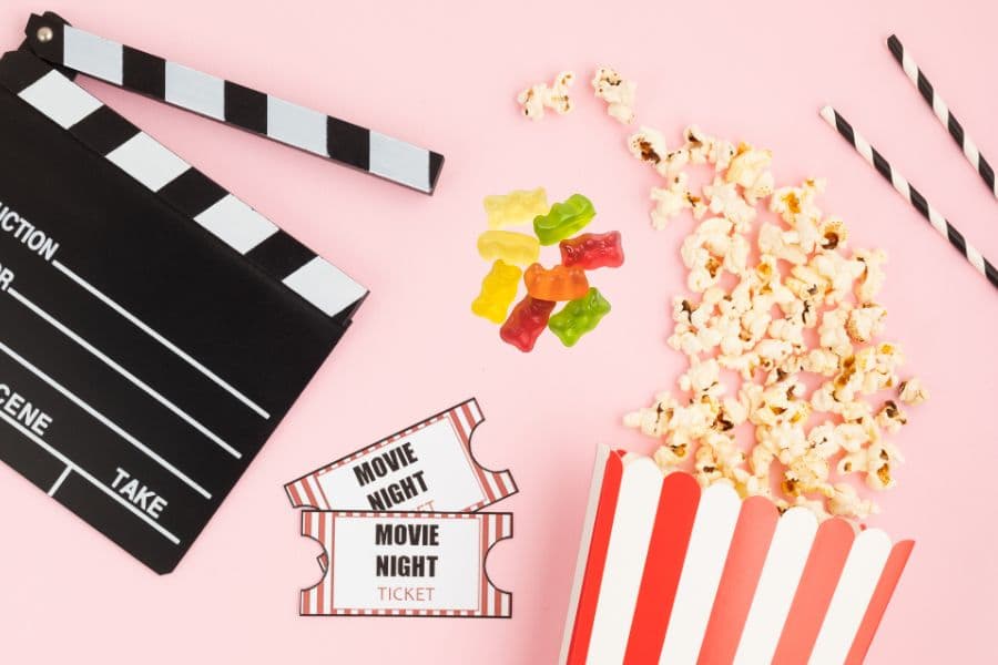 A movie night setup featuring a clapperboard, popcorn, gummy candies, and tickets on a pink background.