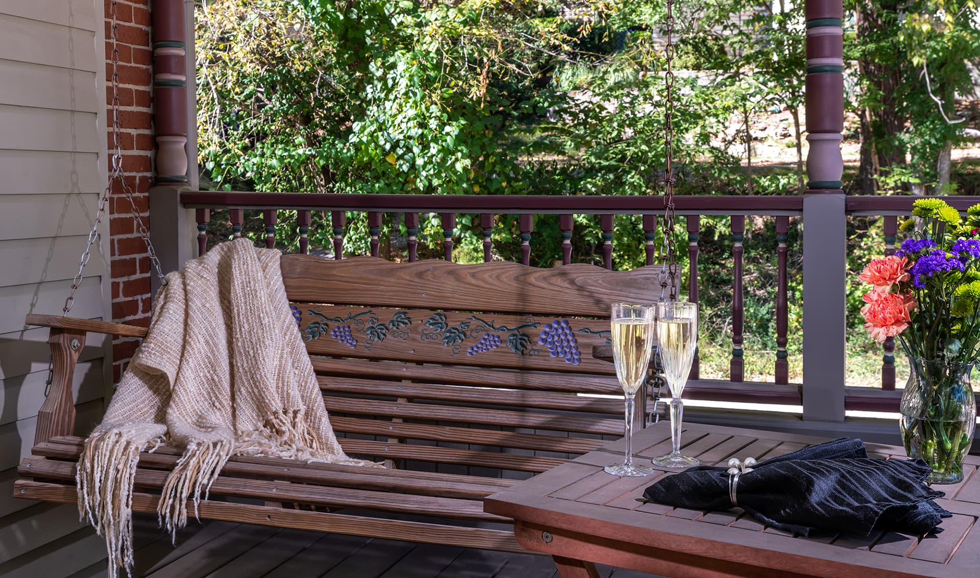 A cozy porch scene featuring a swing, a knitted blanket, two champagne glasses, and a vase of colorful flowers.