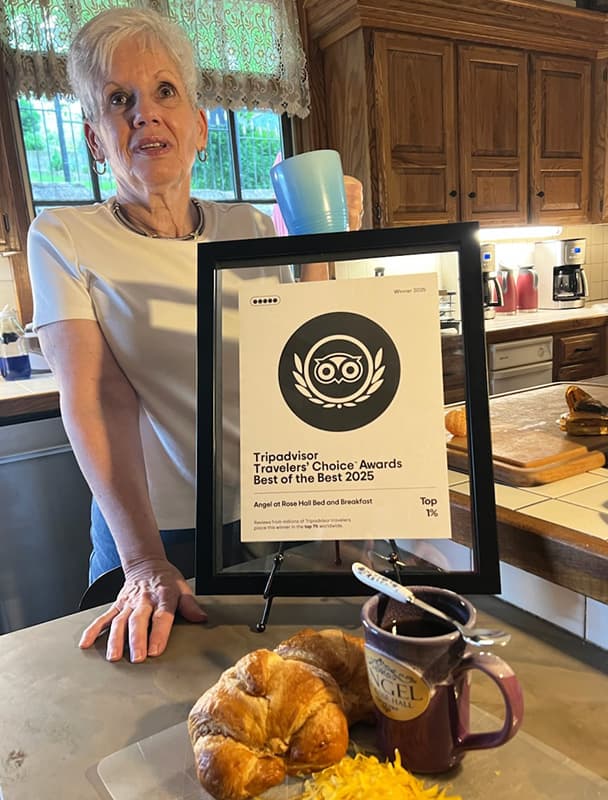 A smiling woman holds a mug beside a framed award for a bed and breakfast, with a croissant and cheese on a plate in the foreground.