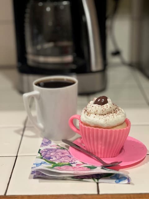 A pink cupcake with whipped cream and a coffee mug sits on a floral napkin beside a silver fork.