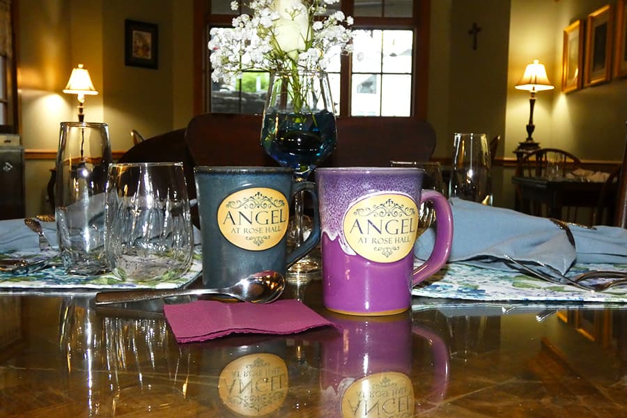 Two mugs labeled "Angel at Rose Hall" sit on a beautifully set dining table with glasses and a small flower arrangement.
