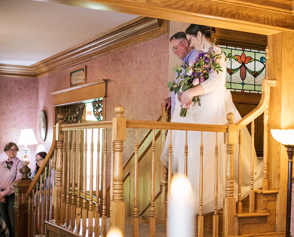 A bride, accompanied by her father, descends a staircase while guests look on.