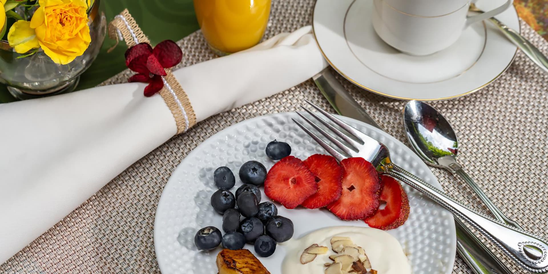 A beautifully arranged breakfast plate featuring yogurt, fresh strawberries, blueberries, and a slice of banana, accompanied by a glass of orange juice and a cup of tea.